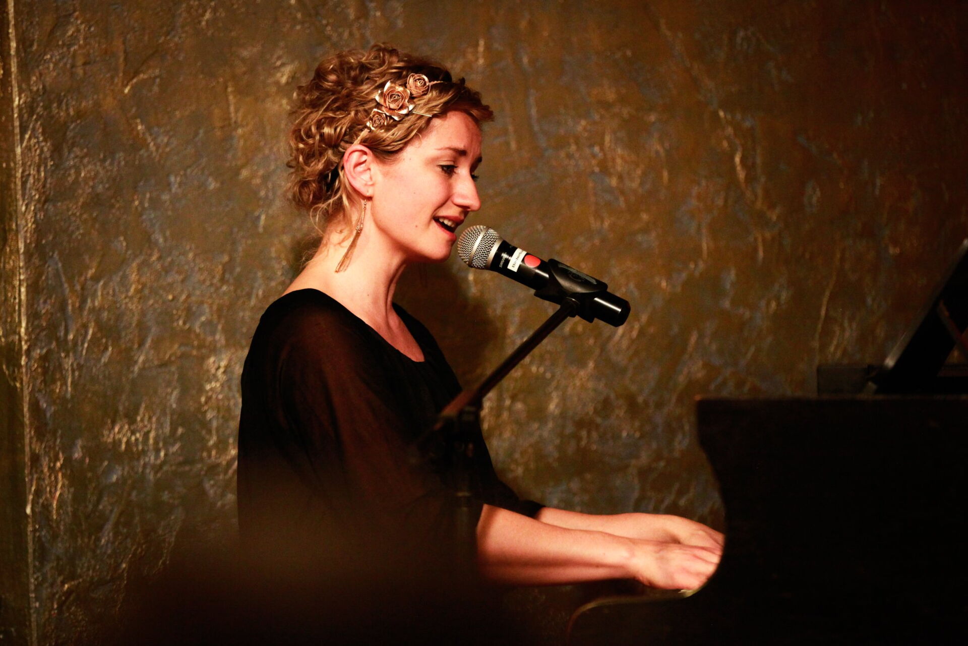 Sängerin mit gelockter Hochsteckfrisur & Mikrofon spielt während dem Singen am Klavier