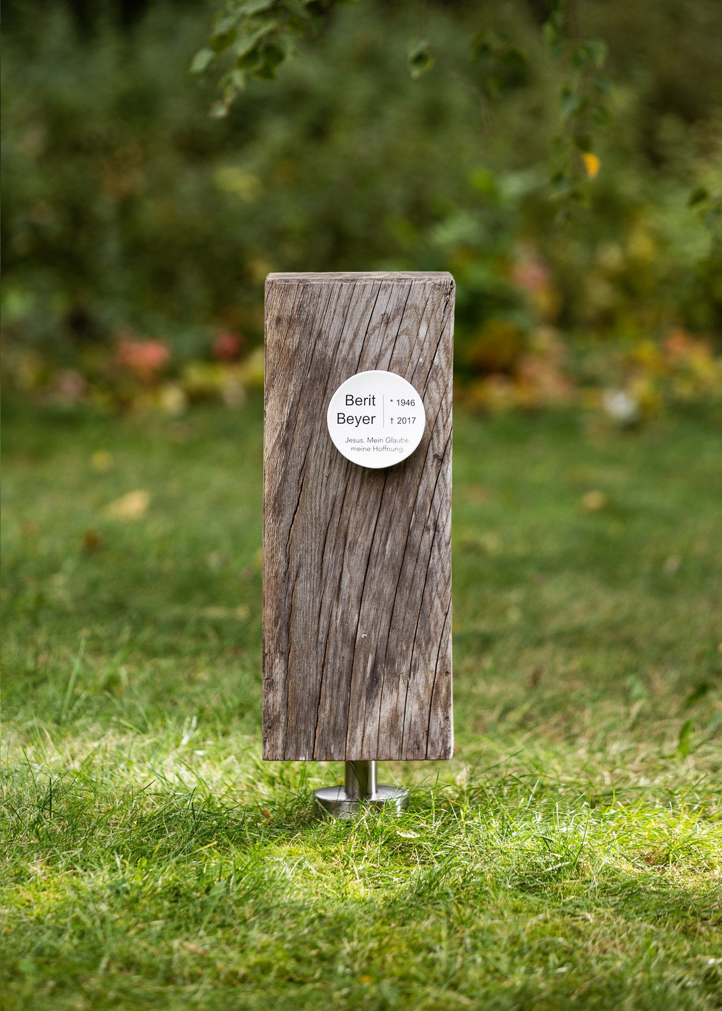 Schlichtes Grabmal aus rechteckigem Holzstück mit kleiner runder Plakette mit Namen der Verstorbenen