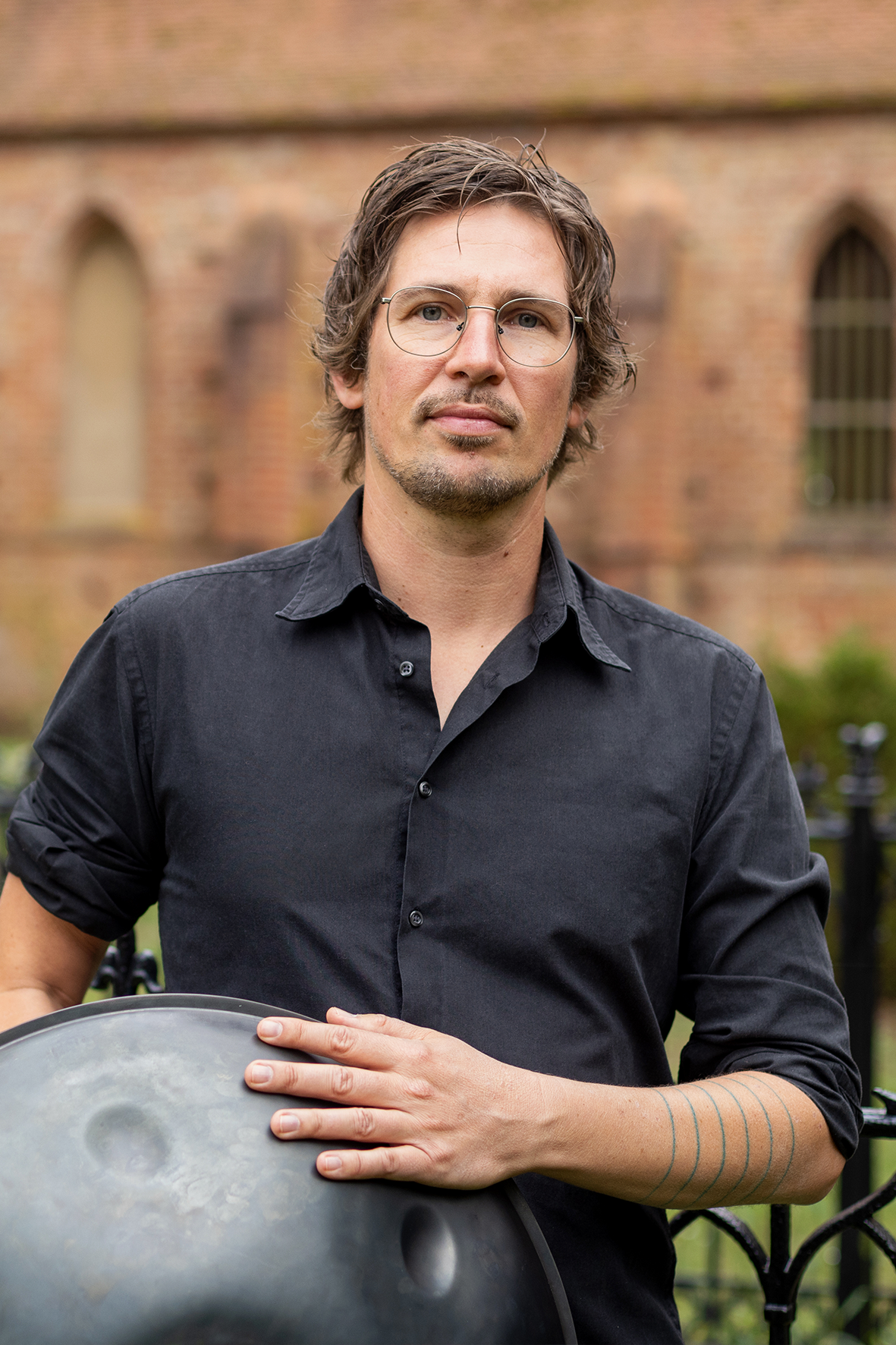 Mann mit dunklem Hemd & Handpan-Trommel, im Hintergrund ein verschwommenes Kirchengebäude